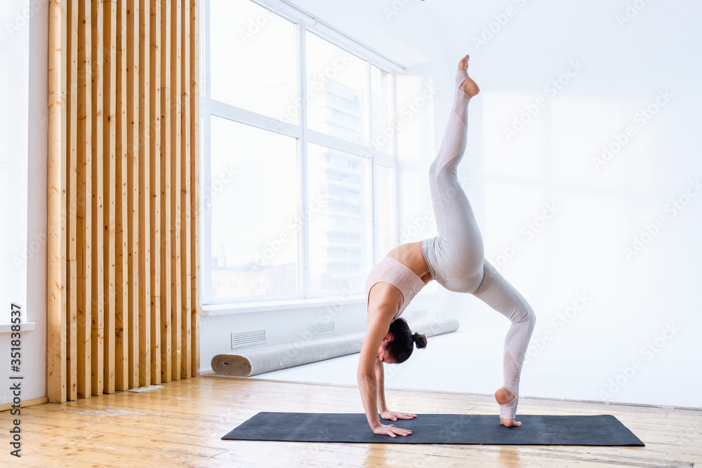 Young caucasian female athlete doing bridge exercise one legged wheel pose while standing on rug on floor of sports gym. Concept of yoga and pilates lovers. Advertising space