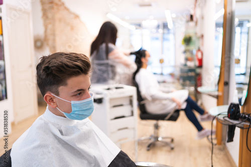 Young boy protected with a mask and keeping a safe distance with another client waiting to have his hair cut at the hairdresser