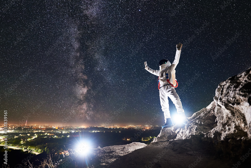 Full length of space traveler raising arms while standing on rocky mountain with fantastic starry sky, milky way on background. Spaceman wearing white space suit and helmet. Concept of space travel