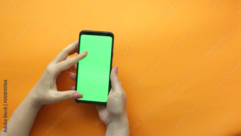 A woman using a smartphone with a green screen on an orange background . Female hands scroll through the pages, tapping on the touch screen. the view from the top. Chroma key. A woman using a smartphone with a green screen on an orange background . Female hands scroll through the pages, tapping on the touch screen. the view from the top. Chroma key.