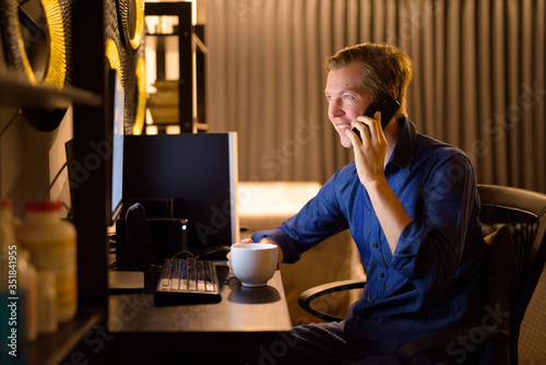 Happy young businessman with coffee talking on the phone while working from home late at night