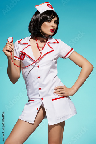 A brunette woman is wearing a white nurse gown with red trimmings and a headband with a red cross symbol. The charming lady is posing on the blue backdrop with a stethoscope in her hand. 
