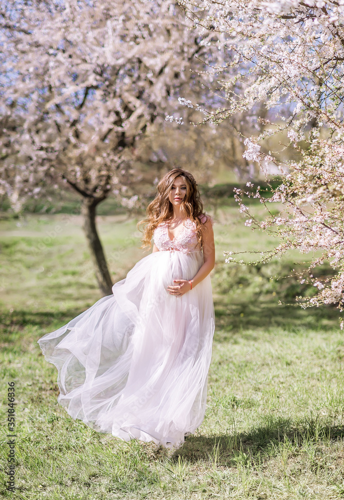 beautiful pregnant woman in a long light dress walks in a blooming spring garden.