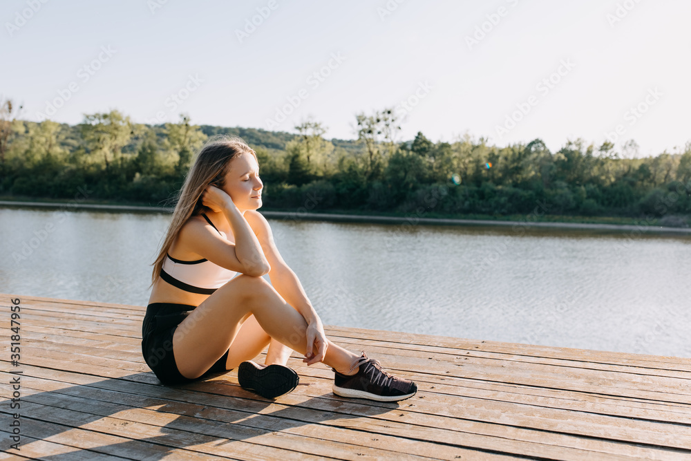 Young slim fit woman resting after workout, sitting on a wooden platform by the lake, outdoors. Sportive girl enjoying sun light and fresh air at the sunrise, smiling.