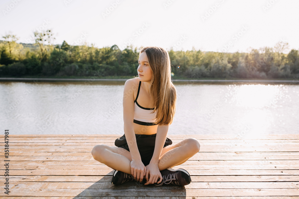 Young slim fit woman resting after workout, sitting on a wooden platform by the lake, outdoors, in the morning.
