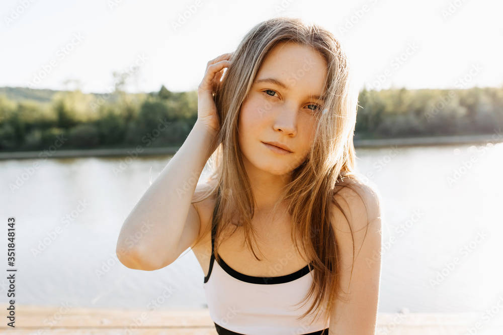 Young blonde woman close-up portrait, sitting by the lake, outdoors, in the sunrise light, looking to camera.