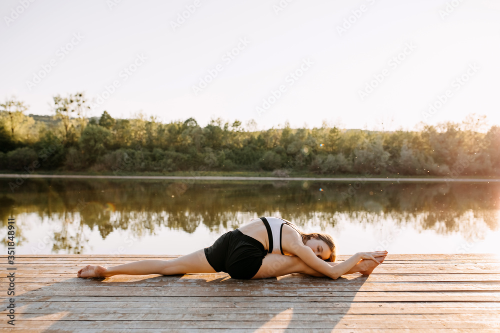 Young slim fit woman doing stretching exercise, twine pose, in the morning. Yoga instructor outdoors, on a wooden platform by the lake.