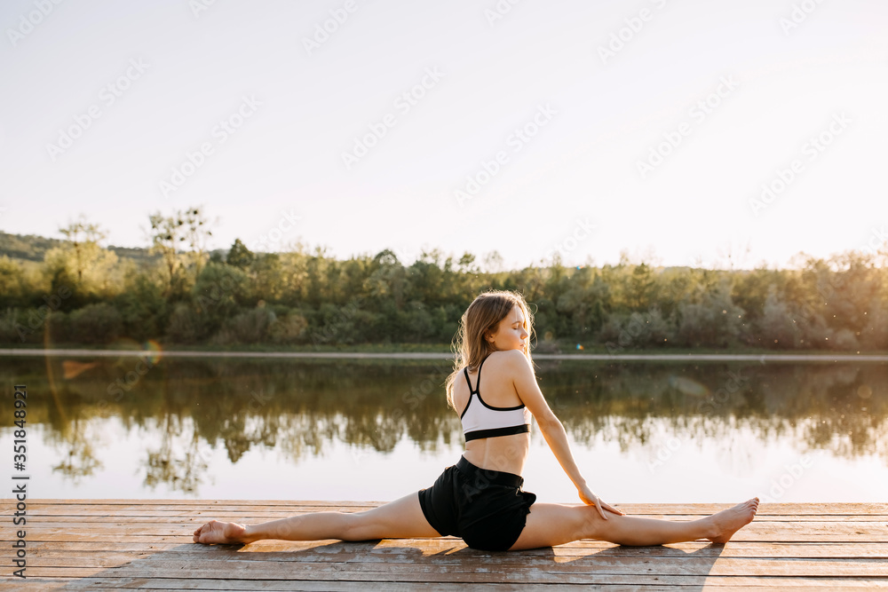 Young slim fit woman doing stretching exercise, twine pose, in the morning. Pilates teacher outdoors, on a wooden platform by the lake.