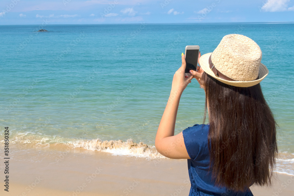 Cute Asian young teenager girl wearing sun hat & navy blue dress using mobile phone or smartphone taking photo or picture of deep blue sea with soft calm waves touching tropical summer sand beach