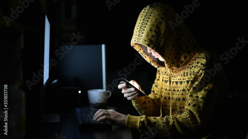 Young man wearing hoodie as hacker using phone and computer in the dark