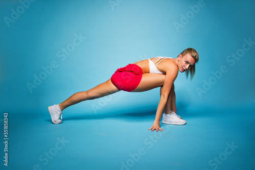 young fitness happy sexy blonde girl in red skirt and white top is doing yoga exercise and looking straight with hands on the floor on the blue wall background, sport concept, free space