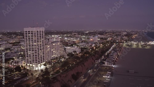 Aerial shot of illuminated buildings and street by beach, drone flying backward over city against sky at night - Santa Monica, California photo