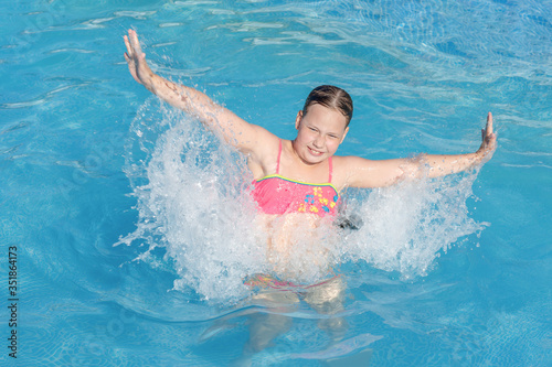 naughty game with water in an outdoor pool at a summer resort. a happy girl in a bright swimsuit jumps in the water with splashes from her hands. Summer vacation and healthy lifestyle concept © Wlad Go