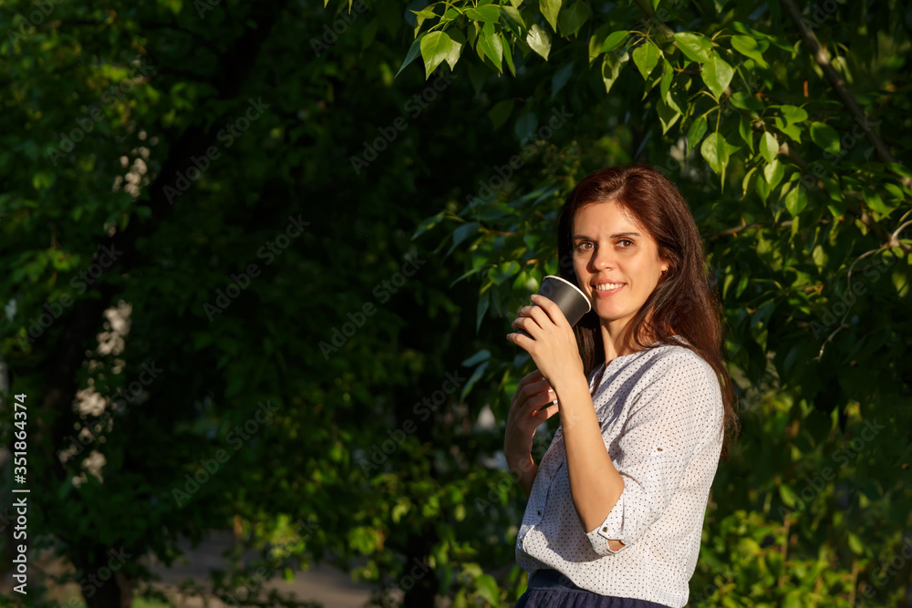 Smiling woman with dark hair holding black paper coffee cup in a garden looks at the camera Smiling woman with dark hair holding black paper coffee cup in a garden looks at the camera