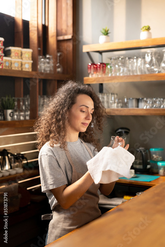 Pretty young waitress in workwear cleaning glass with white cotton napkin
