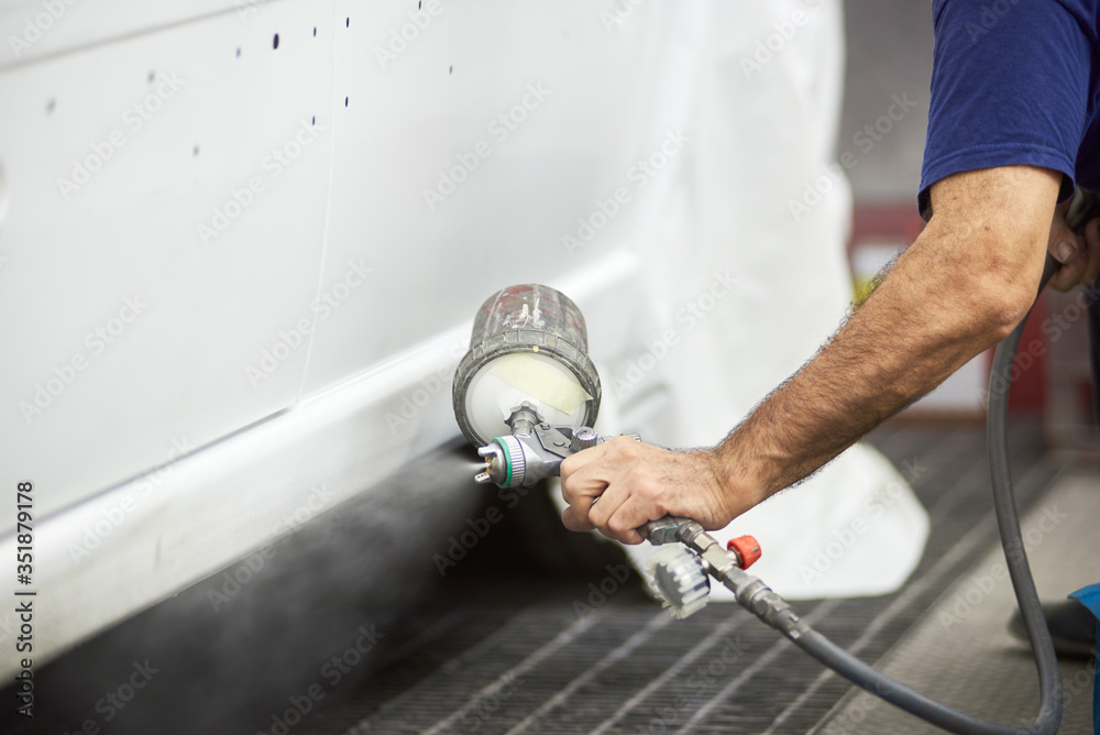Close up of male hand holding spray gun and painting vehicle in white color. Gentleman demonstrating paint spraying technique on automobile. Concept of auto service and tuning.