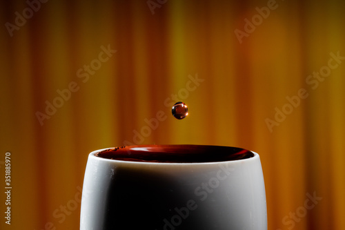 Red wine splash. Reaction of a falling drop on a red wine  surface in a completely filled white plastic cup. Studio photo with matching orange striped colored background. Beautiful wine drop.