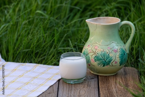 A glass of milk and a jug stand on the table against a background of green grass.