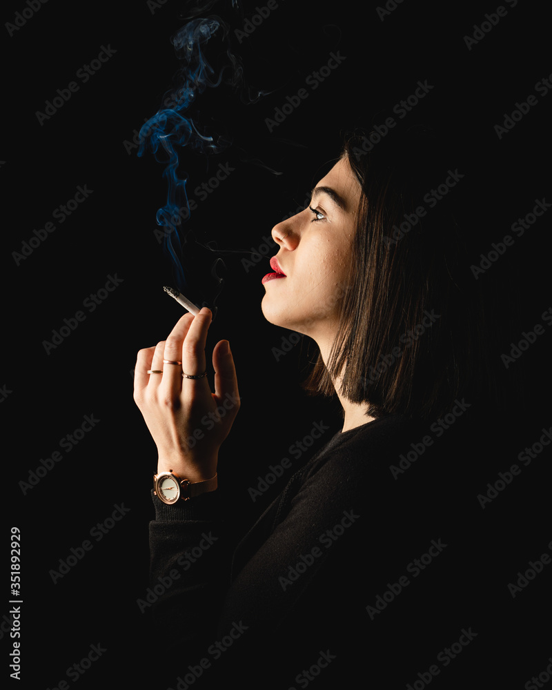 Obraz premium Low key close-up young woman smoking cigarette on black background. 