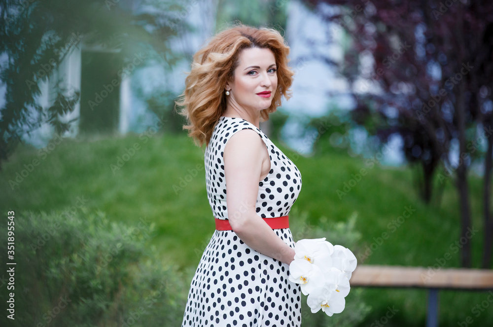 Naklejka premium beautiful red-haired woman in a white dress with black polka dots. Hands with white orchids. Beautiful hair girl. Beauty portrait 