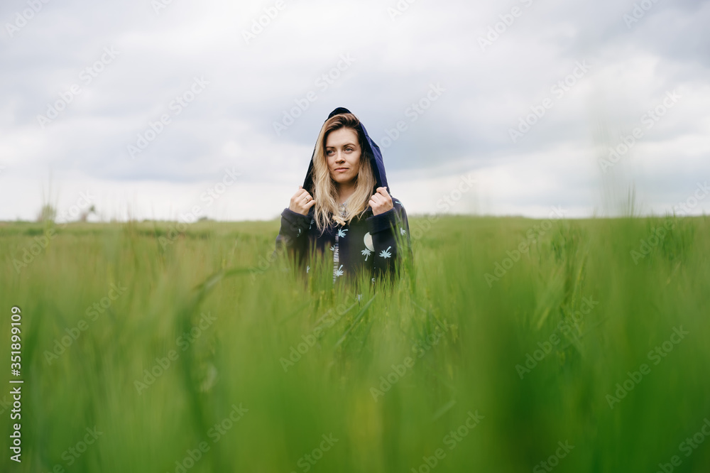 Portrait. .A beautiful girl with long blonde hair in a blue hood is among bright green grass looks into the distance with a smile Portrait. .A beautiful girl with long blonde hair in a blue hood is among bright green grass looks into the distance with a smile