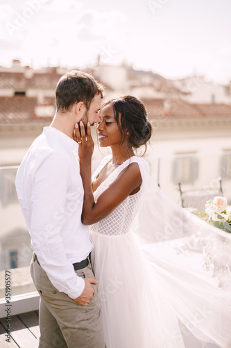 Mixed-race wedding couple. Caucasian groom and African-American bride cuddling on a rooftop in sunset sunlight. Destination fine-art wedding in Florence, Italy. 