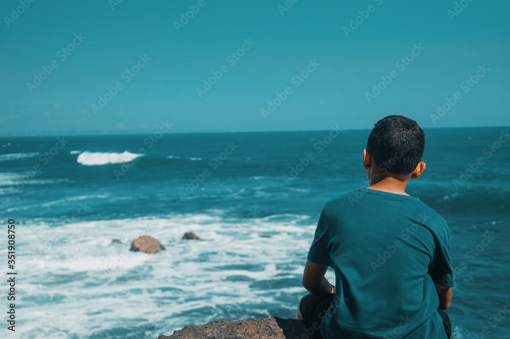 A boy sitting on a rock casually looked out at the ocean and the waves