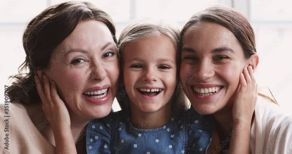 Close up portrait of happy beautiful three female generations family. Head shot little adorable preschool girl embracing smiling middle aged granny and pretty affectionate mixed race mother indoors.