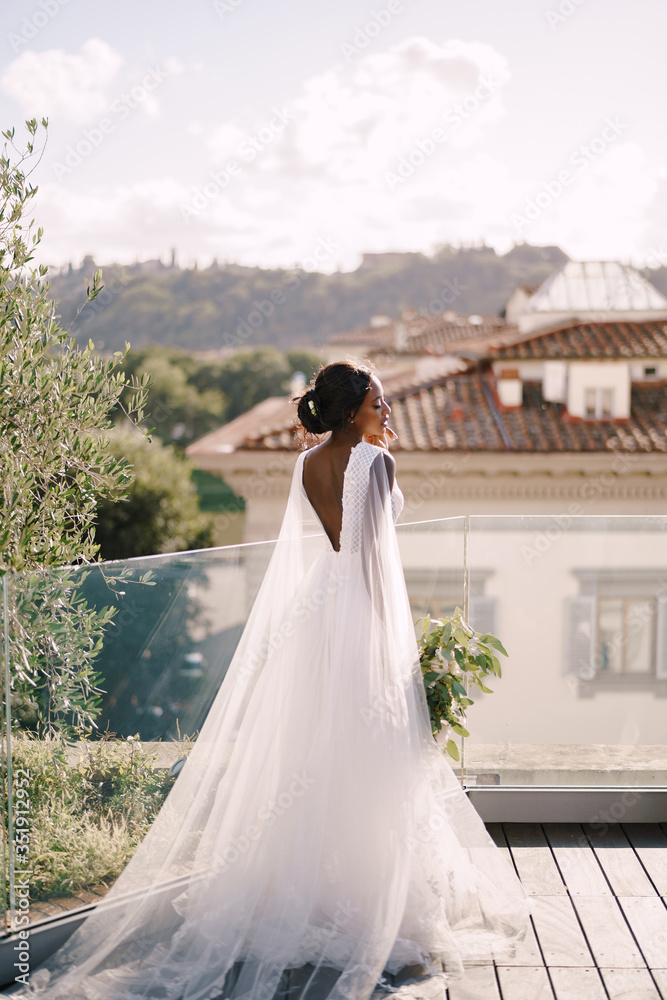 Destination fine-art wedding in Florence, Italy. African-American bride, with a bouquet in her hands, stands on the roof in the sunset light.