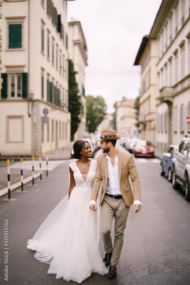 Wedding in Florence, Italy. Multiethnic wedding couple. African-American bride in a white dress and Caucasian groom in a hat are walking along the road among cars.
