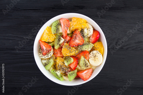 Fruit salad with chia seeds in the white bowl in the center of the black wooden background. Top view. Closeup.
