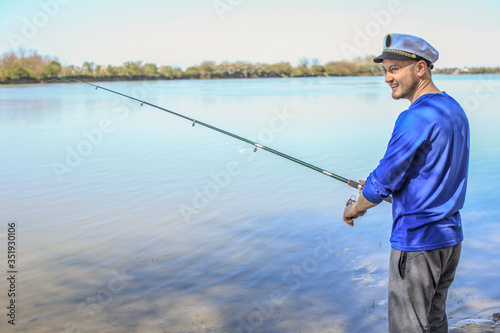 a fisherman in a blue T-shirt and white cap is standing on the river with a fishing rod