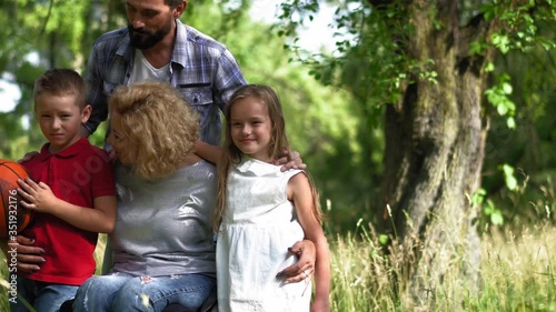 Family with a mother in a wheelchair making a family portrait standing in the nature, children hugging their mother kiss her on the cheek. Family portrait concept. Prores 422. 