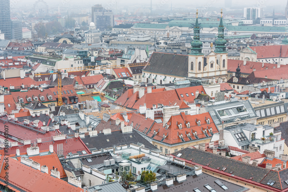 Obraz premium Cityscape with red tiles rooftops and tower of Jesuit Church of the old town of Vienna in a heavy snowy day. View at the tower of St. Stephen's Cathedral in Vienna, Austria.