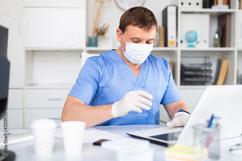 Man doctor in face mask working at laptop