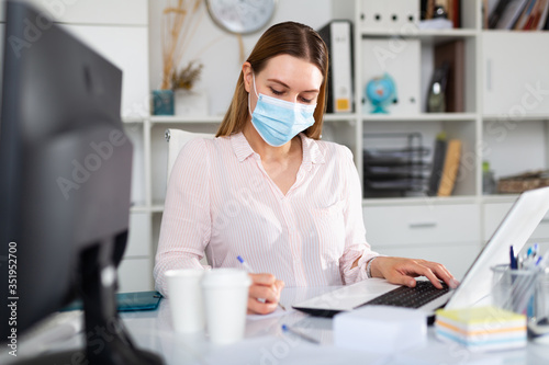 Female office worker in medical mask is having productive day at work in office