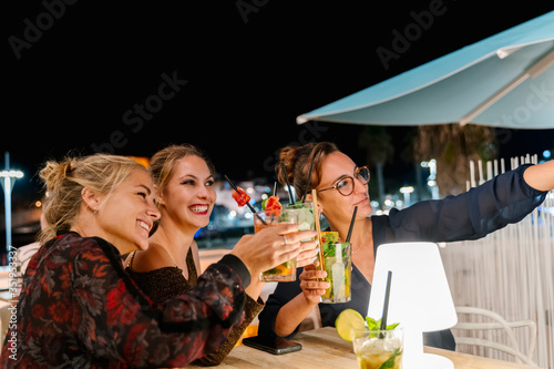 Group of women making a selfie while sitting on a terrace drinking cocktails at night