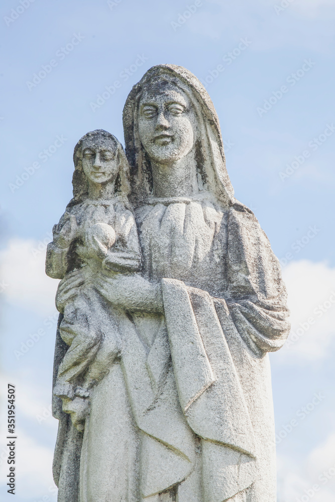 Very ancient stone statue of the Virgin Mary with the baby Jesus Christ against blue sky. Religion, faith, eternal life, God, the soul concept.