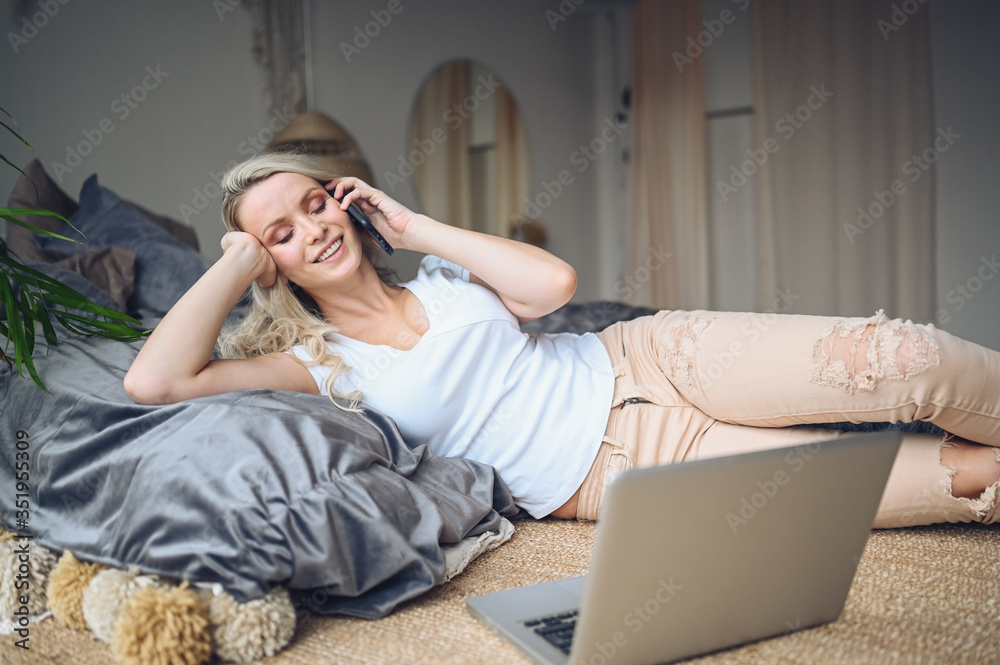 Obraz premium Blonde woman speaking phone working, making video call, watching movie on a laptop in cozy home bedroom modern interior. Home quarantine Covid-19 pandemic Corona virus. Remote work from home concept.