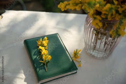 A yellow bouquet in a transparent vase and a green notebook on the table . Still life outdoors on the street. Yellow flowers in a vase in the sun. Green Notepad close-up with sprigs of yellow flowers