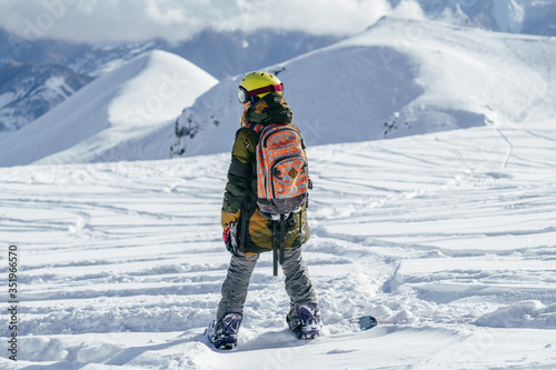 Girl with bright helmet and colourful backpack on a snowboard admires snow-capped mountain peaks. She is choosing the further line for freeride.