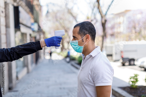 A uomo moro con mascherina chirurgica viene presa la temperatura del corpo con termometro elettronico digitale, isolato su sfondo urbano photo