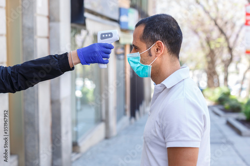 A uomo moro con mascherina chirurgica viene presa la temperatura del corpo con termometro elettronico digitale, isolato su sfondo urbano photo
