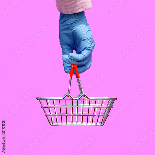A woman's hand in a disposable bright glove holds a shopping metal basket from a supermarket on a lilac background. The safety of customers during the epidemic of coronavirus. Pop art style. photo