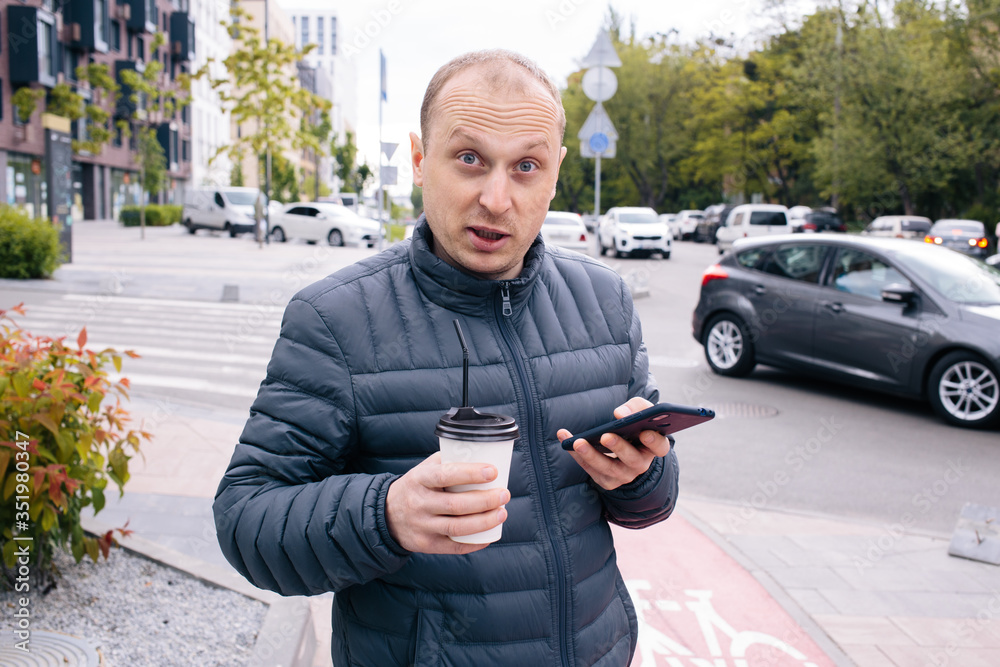 Obraz premium A caucasian positive surprised man in a gray jacket with a white paper cup of Latte coffee or tea with lid and straw and phone on his hand on the city background. Eco. break. Close up