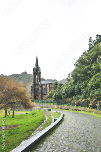 Cobbled path by the lake surrounded by green trees. Chapel of Nossa Senhora das Vitorias in Furnas, Azores Islands, Portugal in the background. Overcast, rainy day, white sky. Vertical photo.