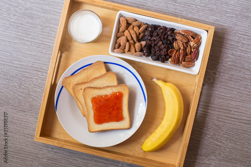 Top view of a meal made with a toast with strawberry marmalade, milk, a banana, almonds, nuts and raisins over a table