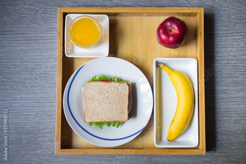 Top view of breakfast made with a ham sandwich, orange juice, a banana and an apple on a wood plate over the table