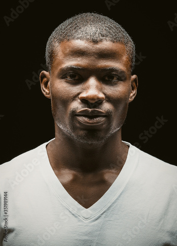 Confident Afro American man thoughtfully looking at camera. Serious attractive dark-skinned guy in a white t-shirt on black background. Toned image
