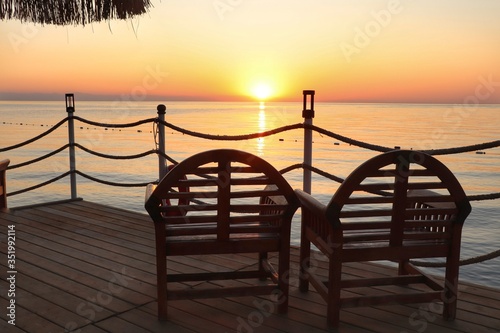 Two chairs stand on the  dock with an orange sunrise over the sea in the background 